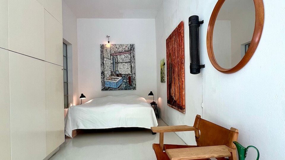 Narrow, minimalist bedroom with white walls and a glossy light floor, featuring a low bed with white bedding at the far end beneath a large framed artwork. Along the right wall are a wooden chair, small round side table with a green glass pitcher, a round wall mirror, and mounted wall art, while the left side has built-in white cabinetry.