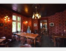 Gemütlicher Raum in einem Restaurant oder Café mit roten, gemusterten Tapeten, Holzstühlen und -tischen sowie warmem Licht aus Wand- und Deckenleuchten. Ein großes Fenster mit Blick auf begrüntes Außenbereich und dekorative Elemente wie ein ausgestelltes Kleid und mehrere Kerzen vervollständigen die Einrichtung.