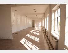 Langer, leerer Innenraum mit weißen Wänden, hellem Holzboden und einer Reihe großer Fenster entlang der rechten Seite. Sonnenlicht fällt durch die Fenster und wirft rechteckige Schatten; unter den Fenstern stehen Heizkörper.