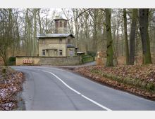 Steinhaus mit Turm, umgeben von einer Mauer, gelegen in einem herbstlichen Waldgebiet; im Vordergrund eine kurvige Landstraße mit weißen Markierungen und leicht bedecktem Boden durch Laub.