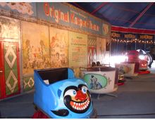 Innenansicht eines Zirkuszelts mit dem "Original Kasperl-Theater". Im Vordergrund eine blau bemalte Karussell-Gondel mit Clownsmotiv, dahinter weitere Gondeln mit dekorativen Illustrationen. Die Wände sind mit bunten Malereien und Reklametafeln verziert.