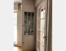 Ein heller Innenraum mit einer weißen Vitrine, die hinter Glasregalen Porzellan und dekorative Gegenstände zeigt. Rechts befinden sich weiße Glastüren, die nach außen führen, und der Boden besteht aus hellem Holz.