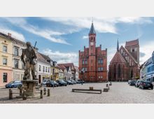 Ein gepflasterter Marktplatz mit einer steinernen Statue im Vordergrund, flankiert von historischen Gebäuden. Im Hintergrund sind ein rotes, backsteingotisches Rathaus mit Uhrturm und eine große Kirche mit Spitzdach sowie gotischen Fenstern zu sehen.