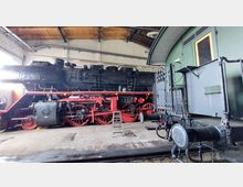 Historische Dampflok in einer Lokhalle, mit schwarzer Lokomotive, roten Rädern und sichtbaren technischen Details. Im Vordergrund ein grüner Waggon und eine Leiter neben der Lokomotive, die unter einem hölzernen Dach abgestellt ist.