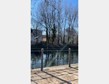 Ein Gehweg mit einem Metallgeländer am Ufer eines Flusses oder Kanals, dahinter eine Baumreihe mit kahlen Ästen. Im Hintergrund sind Gebäude und ein bewachsener Uferbereich zu sehen, bei klarem Himmel und Tageslicht.