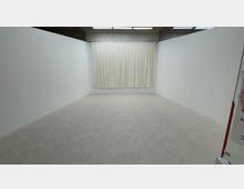 Ein leerer, weiß gestrichener Raum mit glatten Wänden, einem hellgrauen Boden und einer weißen Gardine, die die gesamte hintere Wand bedeckt. Die Decke ist mit rechteckigen Deckenlampen ausgestattet.