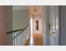 Flur in einem Wohnhaus mit Holzdielenboden, einer weißen Tür im Vordergrund und einem Geländer mit grüner Handlaufleiste. An der Wand hängt ein großer, rechteckiger Spiegel mit goldenem Rahmen, und eine kugelförmige Deckenlampe ist sichtbar.