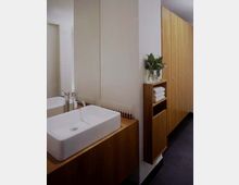 Modernes Badezimmer mit rechteckigem Waschbecken auf einem Holzwaschtisch, großem Spiegel und Schrankelementen aus hellem Holz. Rechts ist ein schmales Regal mit Handtüchern und einer Pflanze zu sehen.