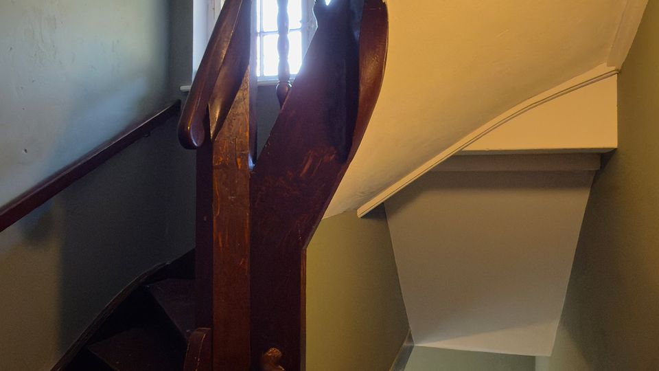 Innenansicht eines schmalen Treppenhauses mit dunkel gebeizter Holztreppe und geschwungenem Geländer, das über mehrere Etagen verläuft. Helle Fenster beleuchten die grünlich gestrichenen Wände, unten steht eine Topfpflanze am Treppenabsatz.