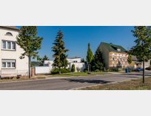 Straßenszene in einem Wohngebiet mit freistehenden Häusern, gepflegten Vorgärten und einer gepflasterten Straße. Auf der linken Seite ein helles Haus mit weißem Putz, rechts ein Gebäude mit gelbem Klinker und grünen Dachziegeln, umgeben von Bäumen und Hecken.