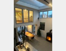 Ein modern eingerichteter Wohnraum mit doppelter Raumhöhe und einer offenen Galerie. Zu sehen sind ein Esstisch aus Holz mit Stühlen, eine hohe Zimmerpflanze, eine beleuchtete Weihnachtsbaumdekoration, ein Kamin mit Feuer sowie große Fenster und ein Oberlicht, die viel Tageslicht einlassen.