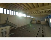 Ein leerstehender Industrie- oder Werkstattraum mit hohen Decken samt sichtbaren Betonbalken. Die Wände sind schlicht und teils abgenutzt, mit mehreren Fenstern in der oberen Hälfte, durch die Sonnenlicht einfällt. Es gibt Treppen zu erhöhten Flächen und vertiefte Aussparungen im Boden.