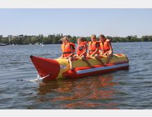 Vier Kinder in orangefarbenen Schwimmwesten sitzen auf einem gelb-roten aufblasbaren Bananenboot, das auf einem ruhigen See schwimmt. Im Hintergrund sind Bäume, einige Gebäude und Boote zu sehen.