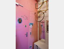 Ein kleiner Raum mit rosa gestrichenen Wänden und einer rosafarbenen Tür, geschmückt mit kleinen Aufklebern und einem runden Fenster. An der Wand hängt ein gelbes Rennrad, daneben befindet sich ein Spiegel, der Teile des Raumes reflektiert, und unten steht ein weißes Möbelstück.