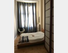 Ein kleines Schlafzimmer mit Holzboden, einem niedrigen Bettgestell aus Holz und einem weißen Bettlaken. Im Hintergrund sind bodenlange, farbige Vorhänge vor einem großen Fenster zu sehen; daneben steht ein geflochtener Stuhl. Rechts im Bild befindet sich ein Raumtrenner mit Holzrahmen und milchigen Paneelen.