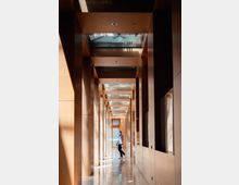 Ein langer, schmaler Korridor mit Holzverkleidungen und großen rechteckigen Fenstern auf beiden Seiten. Die Decke enthält Glasflächen, durch die natürliches Licht einfällt; eine Person geht durch den Korridor.