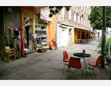 Straßenszene mit einem kleinen pakistanisch-indischen Lebensmittelgeschäft und Imbiss auf der linken Seite, dessen Eingangsbereich von Kleidung und Warenständen gesäumt ist. Rechts daneben befindet sich ein Café mit orangefarbenen Außenwänden und einer Terrasse mit Tischen und Stühlen; im Vordergrund ein schwarzer Tisch mit orangefarbenen Stühlen auf einem Gehweg.
