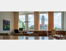 Blick aus einem Büro mit großen Fenstern auf eine städtische Umgebung, im Hintergrund ein historisches Gebäude mit Turm und rotem Dach. Im Vordergrund befinden sich Schreibtische, Stühle und ein abstraktes Gemälde an der Wand.