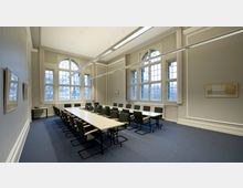 Konferenzraum mit langen, rechteckigen Tischen und gepolsterten Stühlen in einer hellen Umgebung. Der Raum hat große Fenster mit Sprossen, durch die Tageslicht einfällt, sowie eine hohe Decke mit langen, rechteckigen Deckenlampen.