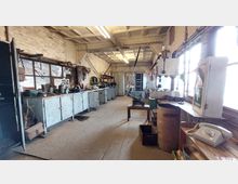 Alter Werkstattraum mit hellen Backsteinwänden, langen Werkbänken, Werkzeugen und Maschinen. Große Fenster auf der rechten Seite lassen Tageslicht in den Raum, der mit Metallregalen, Schränken und alten Geräten ausgestattet ist.