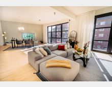 Wohnzimmer in einem modernen Loft mit großen bodentiefen Fenstern, einer grauen Eckcouch mit bunten Kissen, einem Glastisch und heller Holzboden. Im Hintergrund steht ein Essbereich mit schwarzem Tisch und Stühlen sowie einer modernen Dekoration.