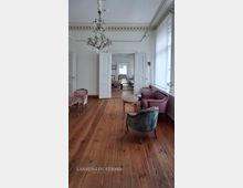 Heller, klassischer Salon in einem Altbau mit hohen Decken, Stuckgesims und großem Kristalllüster über breitem Holzdielenboden. Im Raum stehen mehrere Polstersessel und kleine Beistelltische; doppelflügelige Türen öffnen den Blick in weitere, hintereinanderliegende Zimmer mit großen Fenstern.