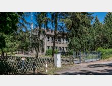 Zweistöckiges, graues Gebäude mit mehreren Fenstern, umgeben von hohen Bäumen und grünem Bewuchs. Im Vordergrund befindet sich ein Holzzaun und ein Metalltor, das den Zugang zur Grundstückseinfahrt markiert.