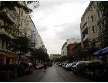 Straßenszene in einer urbanen Umgebung mit mehrstöckigen Wohnhäusern auf beiden Seiten, teils mit Balkonen. Links befinden sich Geschäfte mit Markisen und ein Café mit Außensitzplätzen, während entlang der gepflasterten Straße Autos geparkt sind und einige Bäume stehen.