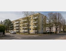 Wohngebiet mit einem mehrstöckigen, gelb verputzten Wohngebäude, das über Balkone an der Vorderseite verfügt. Im Vordergrund befindet sich eine ruhige Straßenkreuzung, umgeben von Bäumen und geparkten Autos.