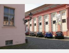 Ein gepflasterter Innenhof mit geparkten Autos, umgeben von historischen Gebäuden mit rosa und cremefarbenen Fassaden, klassizistischen Säulen und großen Fenstern. Eine schmiedeeiserne Laterne ist an der Hauswand befestigt, und ein Banner hängt an der Fassade rechts.