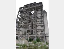 Altes, verfallenes Industriegebäude mit Betonrahmenstruktur und großen zylindrischen Silos, umgeben von Vegetation und stehenden Wasserflächen. Die Fassade zeigt Verwitterungsspuren, und teils sind Treppen und Plattformen sichtbar.