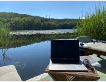 Ein See mit ruhiger Wasseroberfläche, umgeben von dichtem Wald, und einem Schilfgürtel im Vordergrund. Auf einer hölzernen Terrasse befinden sich ein Laptop, eine Sonnenbrille und eine Liegestuhlfläche, die auf eine entspannte Arbeitsumgebung im Freien hindeuten.