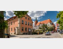 Straßenszene in einer Altstadt mit einem orangefarbenen Gebäude im Vordergrund, einem kleinen Café mit Sonnenschirmen und Fahrrädern daneben sowie einer Kirche mit rotem Dach und spitzem Turm im Hintergrund. Bäume und geparkte Autos säumen die gepflasterte Straße.