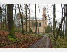 Sandstein-Kirche mit Glockenturm und grünlicher Kuppel, umgeben von einem Wald mit kahlen Bäumen im Herbst oder Winter; ein unbefestigter Weg führt durch das bewaldete Gelände zur Kirche.