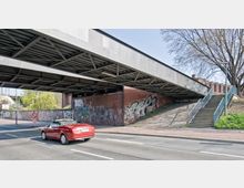 Eine Straßenunterführung mit einer Betonbrücke, darunter eine Backsteinmauer mit Graffiti. Rechts führen Treppenstufen mit einem Metallgeländer zu einem höher gelegenen Bereich, während links ein rotes Auto auf der Straße fährt.