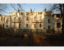 Ein historisches Gebäude mit heller Fassade, großen Fenstern und kleinen Balkonen. Im Vordergrund sind kahle Bäume und ein naturbelassener, bewaldeter Bereich sichtbar.