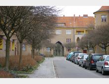 Wohnsiedlung mit mehrstöckigen, sandfarbenen Gebäuden, orangefarbenen Ziegeldächern und gelben Fensterrahmen. Eine gepflasterte Straße mit geparkten Autos wird von kahlen Bäumen und schmalen Grünflächen gesäumt.