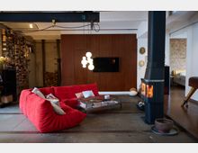 Wohnzimmer in einem industriellen Stil mit Betonboden und freiliegender Decke. Zu sehen sind ein rotes, gepolstertes Sofa, ein Glastisch mit Büchern, ein Kaminofen, eine Wandverkleidung aus Holz, eine Hängelampe mit kugelförmigen Leuchten und Regale voller Bücher im Hintergrund.