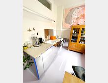 Kleine Küche mit einer Arbeitsplatte, auf der Haushaltsgeräte wie eine Kaffeemaschine und Küchenutensilien stehen. Im Hintergrund befindet sich ein Schrank aus Holz und ein großes abstraktes Gemälde an der Wand.