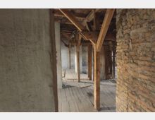 Holzstruktur eines alten Gebäudes mit sichtbaren Holzbalken, einem Holzboden und Wänden aus rohen Ziegelsteinen. Die Umgebung ist unrenoviert und wirkt verlassen.