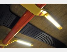 Decke mit sichtbaren Betonplatten, roten und gelben Rohrstrukturen und rechteckigen, in Gelb eingefassten Leuchtstofflampen. Schwarze Lüftungselemente verlaufen parallel zur Beleuchtung.