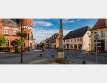 Städtischer Platz mit einem roten Backsteingebäude mit Turm links, einer dekorativen Säule in der Mitte und umliegenden Fachwerk- und Wohnhäusern. Der Platz ist von Bäumen und Laternen umgeben, und im Hintergrund erstreckt sich eine gepflasterte Straße mit historischen Gebäuden.
