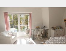 Helles Wohnzimmer mit großen Fenstern und Blick auf einen Garten, ausgestattet mit einem weißen Sofa, einem Glastisch mit Holzstühlen sowie einer Kommode und Schreibtisch auf der rechten Seite; rosa Vorhänge rahmen die Fenster ein.