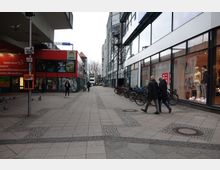 Straßenszene in einer Einkaufsgegend mit breitem Gehweg, links ein rotes Gebäude mit Schaufenstern und Werbung, rechts ein modernes Gebäude mit großen Glasfassaden und Schaufenstern. Fahrräder sind entlang der Gebäude abgestellt, und mehrere Fußgänger sind zu sehen.