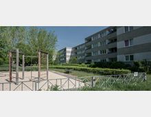 Spielplatz mit Klettergerüst aus Holz und Netz auf einer Sandfläche, umgeben von niedrigen Wohnblöcken mit grauen Fassaden. Im Hintergrund befinden sich Grünflächen, Bäume und Büsche.