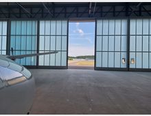 Ein Flugzeughangar mit offenen, teils durchsichtigen Schiebetoren, die den Blick auf eine Start- und Landebahn und eine grasbewachsene Umgebung freigeben. Im Vordergrund ist der Flügel und ein Teil des Rumpfes eines Flugzeugs sichtbar.