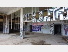 Abgeblendeter Innenraum eines verlassenen Gebäudes mit graffitibeschmierten Wänden, zerbrochenen Fensterscheiben und teilweise zerstörtem Bodenbelag. Die weiß gefliesten Wände und offenen Türöffnungen verstärken den Eindruck von Verfall.