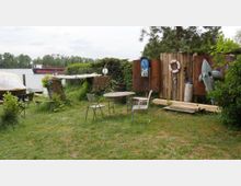 Grüne Uferlandschaft mit einem einfachen Sitzbereich aus Metalltischen und -stühlen, umgeben von Büschen und Bäumen. Im Hintergrund sind ein Fluss, ein großes Schiff und verschiedene Dekorationsgegenstände wie ein Rettungsring, Holzplanken und ein aufblasbarer Hai zu sehen.