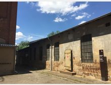 Backstein- und Betongebäude eines historischen Industrieareals mit hohen rechteckigen Fenstern und rostfarbener Patina, flankiert von einem schmalen Hof. Im Vordergrund ein Schild mit der Aufschrift „Mechanische Werkstatt“, daneben eine alte Metalltür.