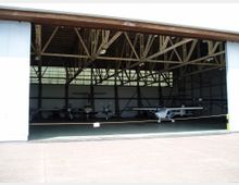 Ein offener Hangar mit mehreren kleinen Propellerflugzeugen im Inneren. Die Dachkonstruktion besteht aus freiliegenden Holzträgern, und das Gebäude hat große Fensterflächen im oberen Bereich, die für Licht sorgen.